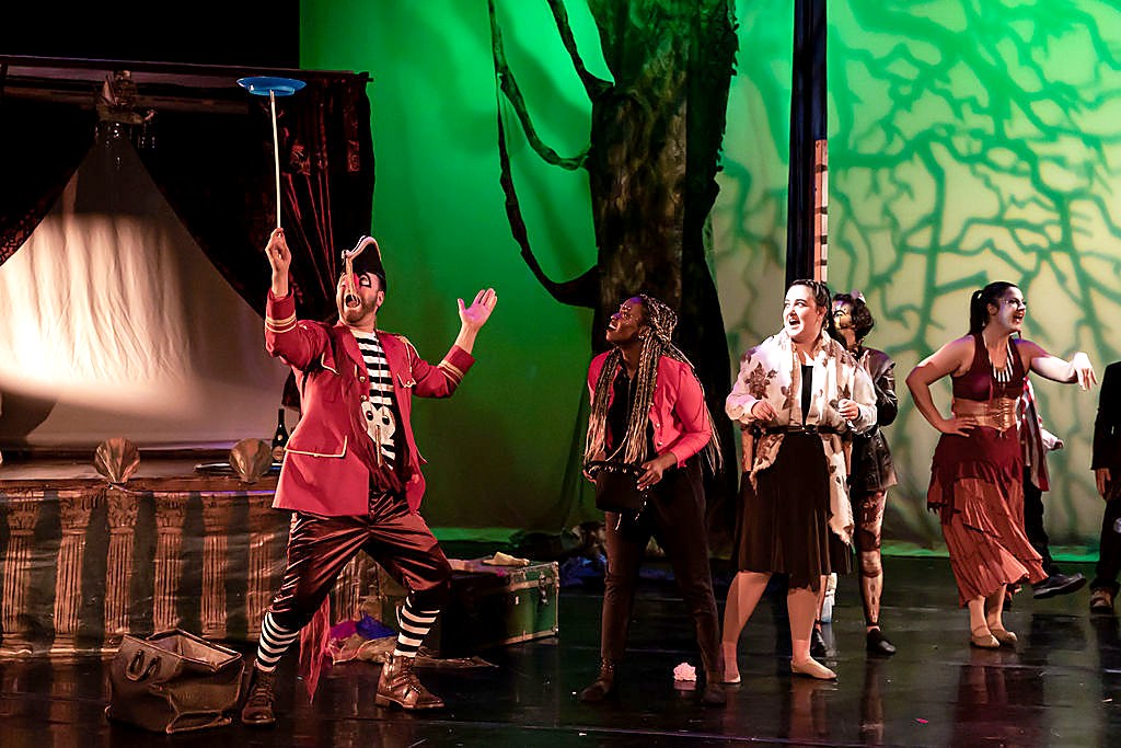 Ensemble cast performing a plate-spinning bit on a forest stage set